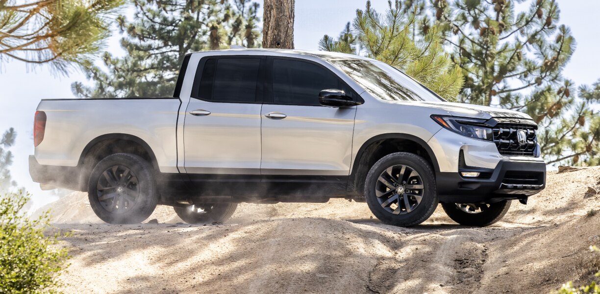 Side wide view of grey Ridgeline driving in a wooded area. 