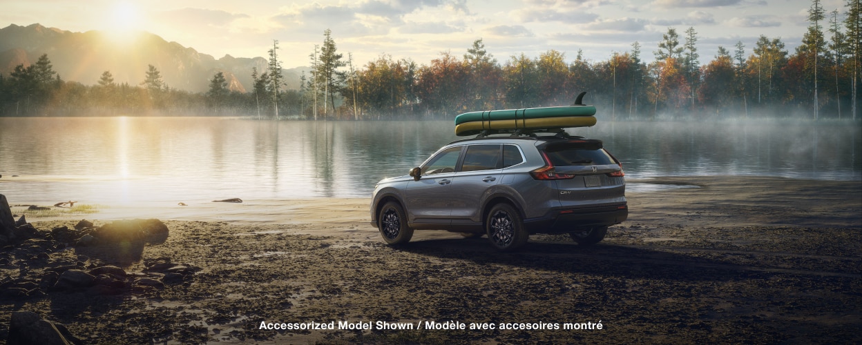 Vue latérale arrière 3/4 d’un CR-V gris, avec deux planches à pagaie sur la galerie de toit, stationné sur le bord d’un lac au petit matin.