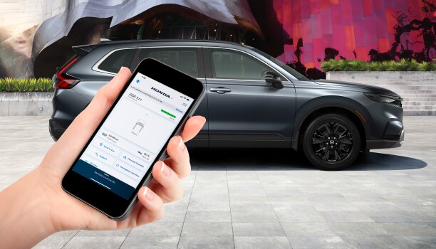 Sideview of grey CR-V parked in front of a graffitied wall. Hand in foreground holds a smartphone with the HondaLink™ app. 