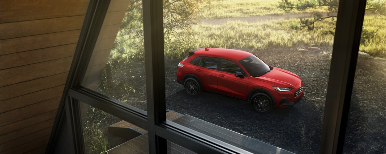 Vue 3/4 en plongée d’un HR-V rouge stationné à la campagne, vu par les fenêtres d’un chalet en A.