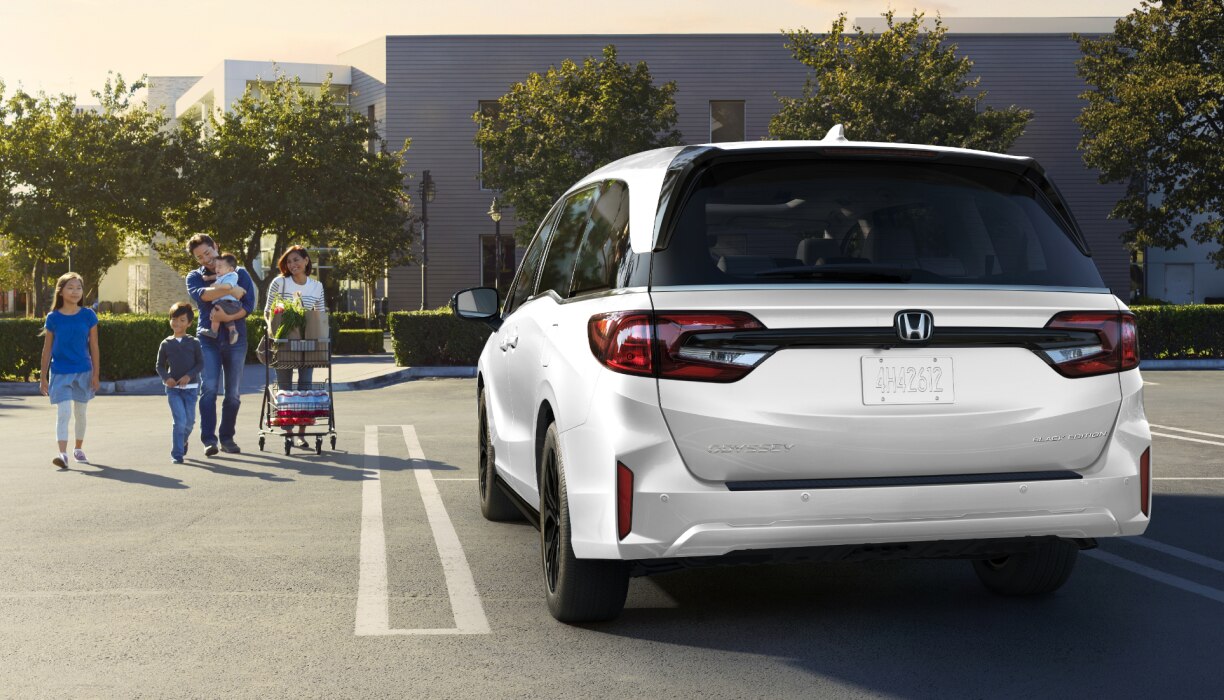 Rear view of grey Odyssey in empty parking lot. Family of walks toward it, pushing a grocery cart.