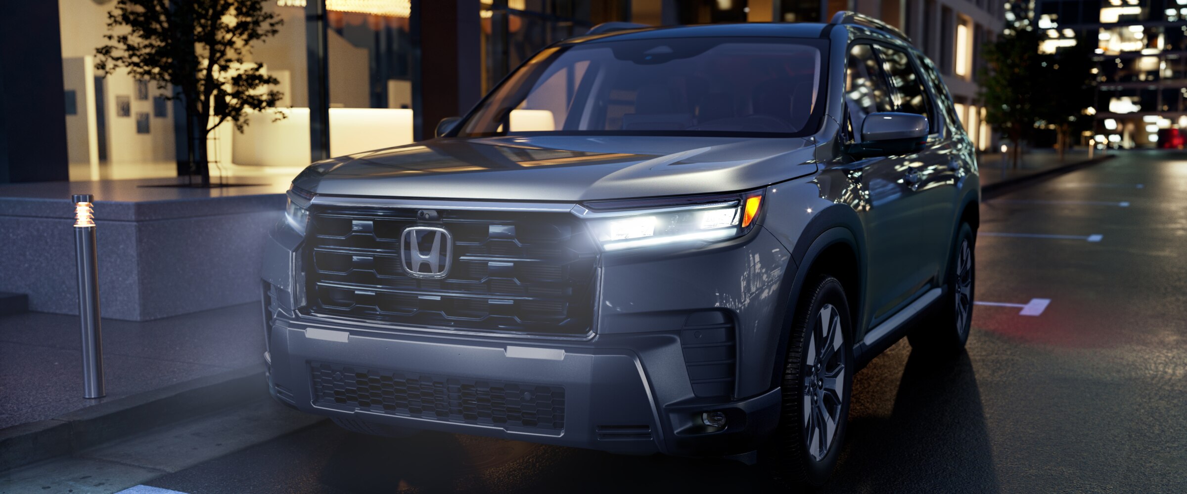 3/4 front side view of a silver Pilot parked on a city street with headlights illuminated. 