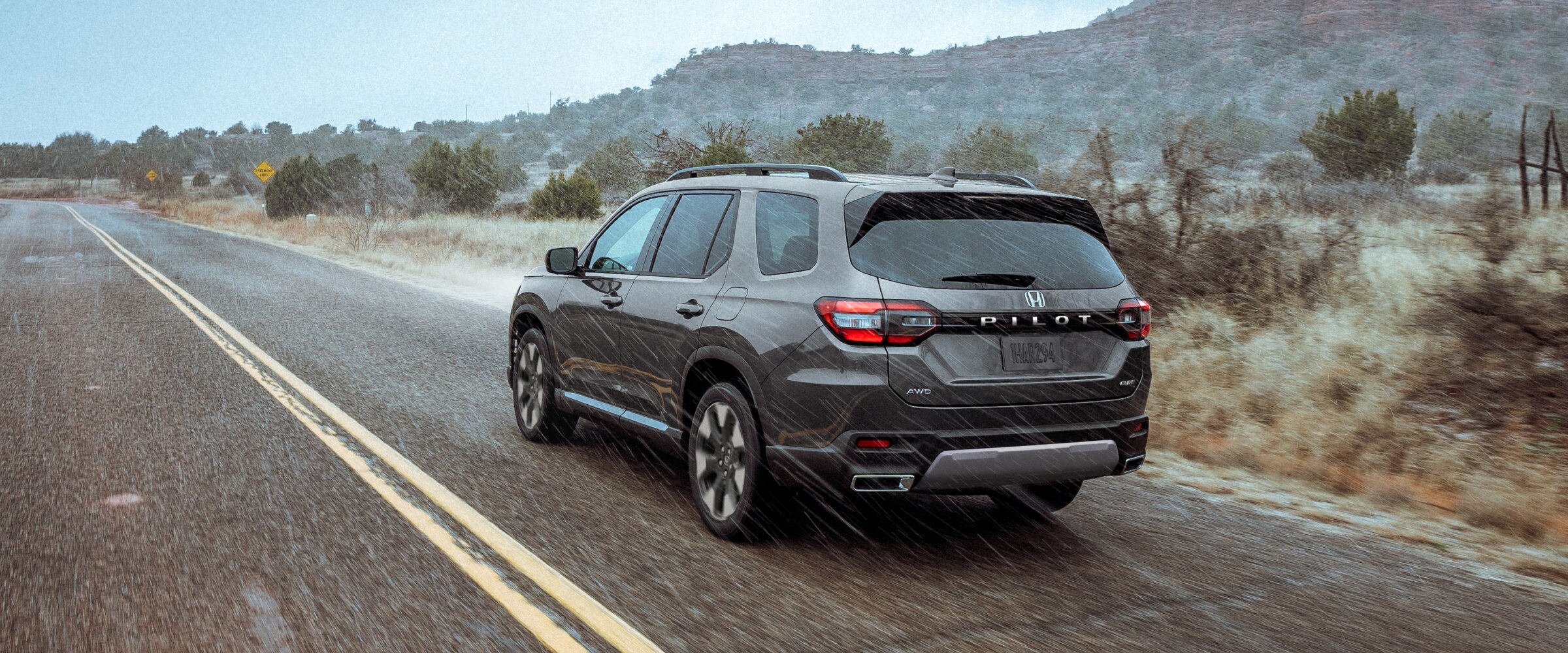 3/4 rear view of a grey Pilot driving through the rain on a highway set against a rugged landscape of plants and brush.