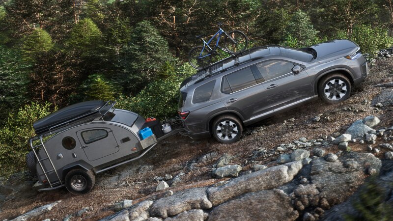Side view of a grey Pilot driving uphill over rocky terrain, with a bike mounted on the roof and a trailer hitched up to the rear.