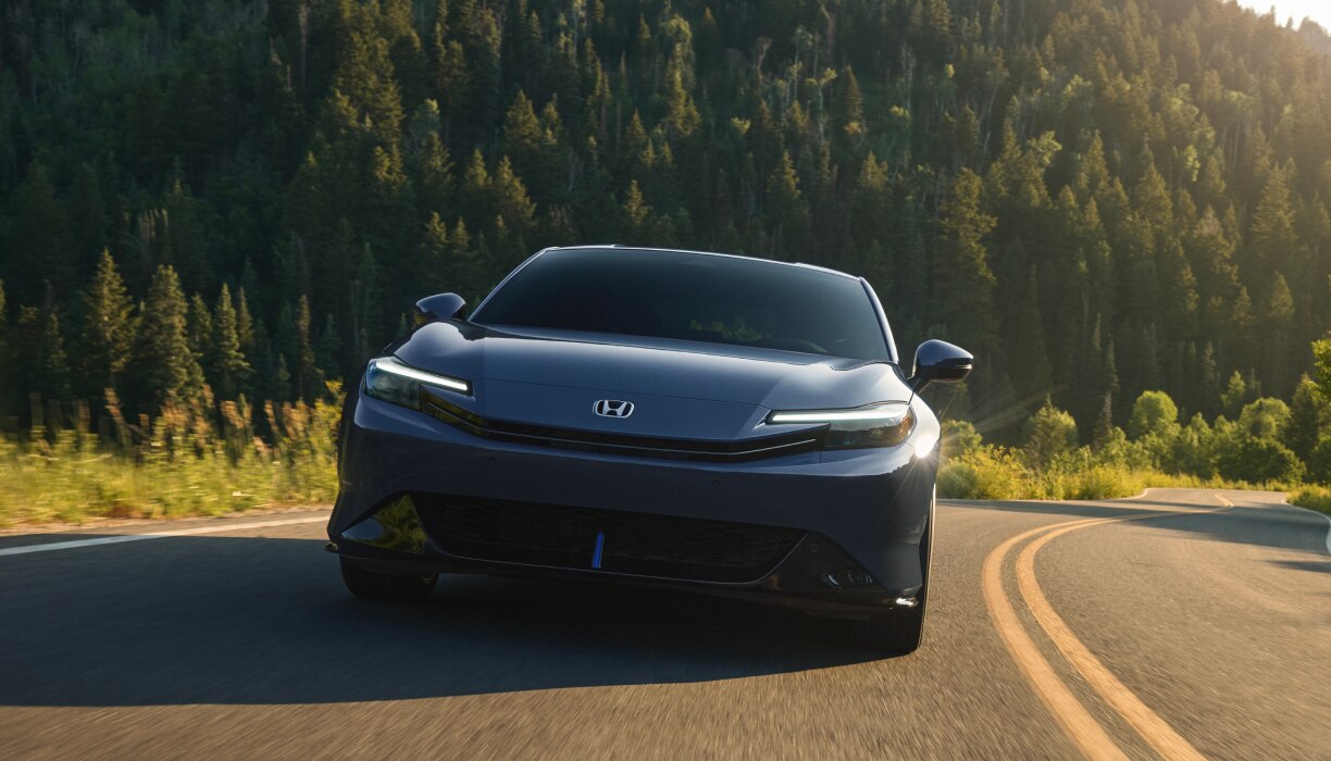 Front view of a Black Prelude driving on a highway framed by forest and warm light. 