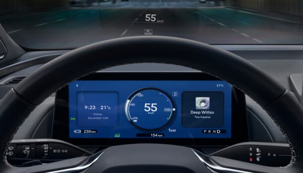 Closeup of instrument cluster screen seen through the steering wheel, indicating info, such as: time, outside temperature, speed, battery charge, and odometer.