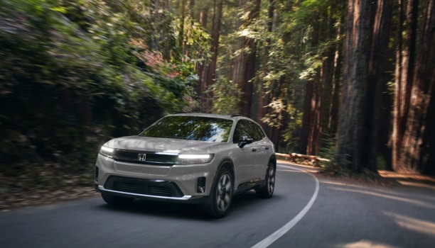 A grey Honda Prologue drives on a road surrounded by trees.