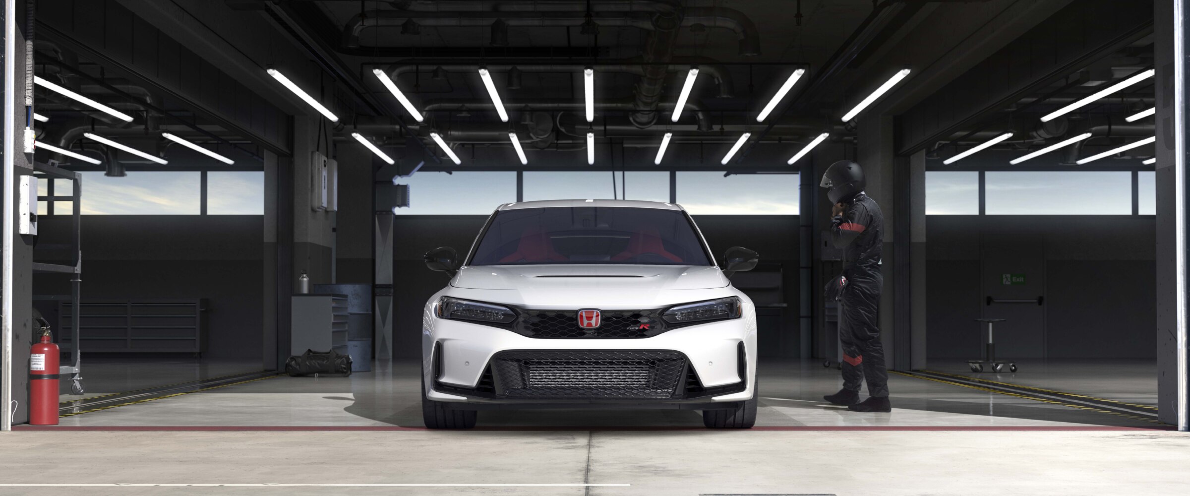 Front view of a white Type R parked in racing garage. A race car driver in black coveralls and helmet nears it. 